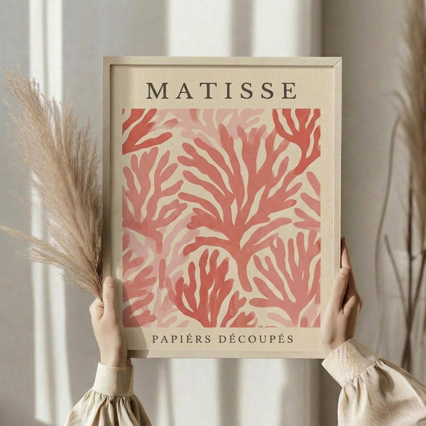 May include: Hands holding a framed art print featuring coral-pink abstract cut-out shapes in Matisse's papier découpé style on cream background, with "MATISSE" and "PAPIERS DÉCOUPÉS" text. Light wood frame, person wearing beige ruffled sleeves, dried pampas grass visible, neutral curtained background with soft natural lighting.