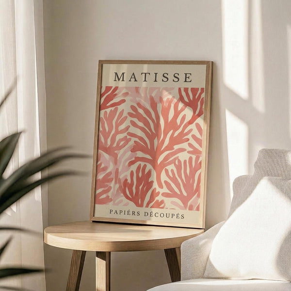 May include: Framed art print with "MATISSE" and "PAPIERS DÉCOUPÉS" text above and below coral pink abstract cut-out pattern on cream background, displayed on round wooden side table next to beige upholstered chair with plant leaves visible in soft natural lighting against white wall.