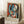 May include: Framed Picasso-style cubist poster featuring abstract woman with blue and white geometric face, black hair, set against colorful background of reds, blues, and yellows. Light wooden frame leans against white wall on textured rug, beside wooden side table with white coffee cup and beige throw blanket.