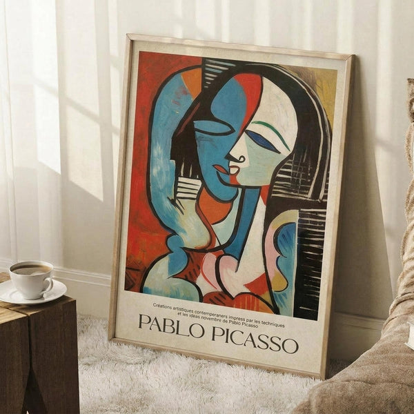 May include: Framed Picasso-style cubist poster featuring abstract woman with blue and white geometric face, black hair, set against colorful background of reds, blues, and yellows. Light wooden frame leans against white wall on textured rug, beside wooden side table with white coffee cup and beige throw blanket.
