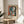 May include: Framed Pablo Picasso exhibition poster featuring abstract portrait of woman with blue and white face, red lips, black hair, in cubist style with bold red, blue, and cream colors. Light wood frame sits on round wooden side table next to beige upholstered chair. Soft natural lighting creates shadows on white wall, with green plant leaves visible in corner of minimalist modern room setting.