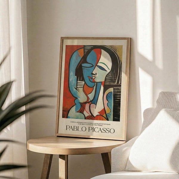 May include: Framed Pablo Picasso exhibition poster featuring abstract portrait of woman with blue and white face, red lips, black hair, in cubist style with bold red, blue, and cream colors. Light wood frame sits on round wooden side table next to beige upholstered chair. Soft natural lighting creates shadows on white wall, with green plant leaves visible in corner of minimalist modern room setting.