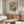 May include: Pablo Picasso exhibition poster featuring cubist artwork of two seated figures in blue, orange and white tones, displayed in thin frame above walnut credenza. Modern living room with cream sculptural chair, glass oval coffee table, white mushroom lamp, and brass floor mirror. Neutral walls with natural lighting.