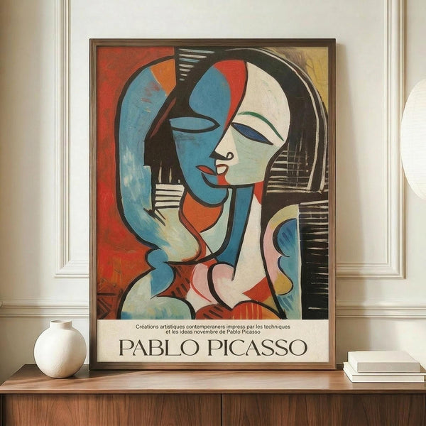 May include: Framed Picasso-style cubist poster featuring abstract woman with geometric blue and white face, red lips, black hair, displayed on wooden sideboard next to white ceramic vase and books in elegant room with white paneled walls