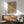 May include: Vintage-style botanical poster with earth-toned leaves, plants and sun motifs in a wooden frame, displayed above a mid-century modern credenza with table lamp. Teal upholstered chair with wooden arms sits nearby on wooden flooring. Natural sunlight creates geometric shadows on white wall, creating a warm, retro-inspired living room scene.