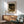 May include: Framed Claude Monet exhibition art print hanging on white wall above wooden sideboard with table lamp. Green upholstered armchair with wooden frame sits nearby. Artwork shows woman in white dress with parasol walking through garden with flowering bushes. Natural sunlight creates dramatic shadows across the mid-century modern furniture and warm wood tones.