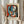 May include: Person in cream sweater holding framed Pablo Picasso poster print featuring abstract cubist portrait of woman with blue and red geometric forms, black hair, against colorful background. Light wood frame with cream matting. Dried pampas grass visible on left side, soft natural lighting from window.