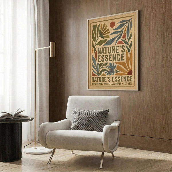 May include: Framed vintage-style botanical poster with green leaves, orange sun, and "Nature's Essence" text in brown lettering, displayed above a beige upholstered armchair with geometric patterned pillow. Modern living room setting features wood paneled wall, brass floor lamp, small round side table with open book, and natural lighting from window with curtains.