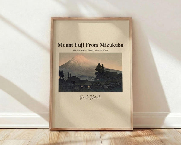 May include: Framed vintage Japanese woodblock print showing Mount Fuji at sunset with pink-tinted peak rising above dark silhouetted hills and traditional buildings. The artwork features muted earth tones in a classic ukiyo-e style, displayed in a light wood frame against a cream textured wall with natural lighting creating soft shadows on wooden flooring.