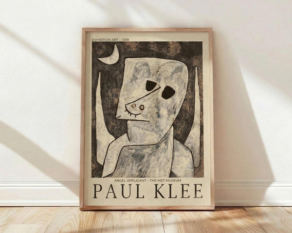 May include: Framed exhibition poster featuring Paul Klee's "Angel Applicant" artwork from 1939, showing an abstract white figure with angular head against dark background under crescent moon. Poster has cream-colored border with black text identifying the artwork and The Met Museum. Natural wood frame sits on light wooden floor against white wall with soft window light casting diagonal shadows across the scene.