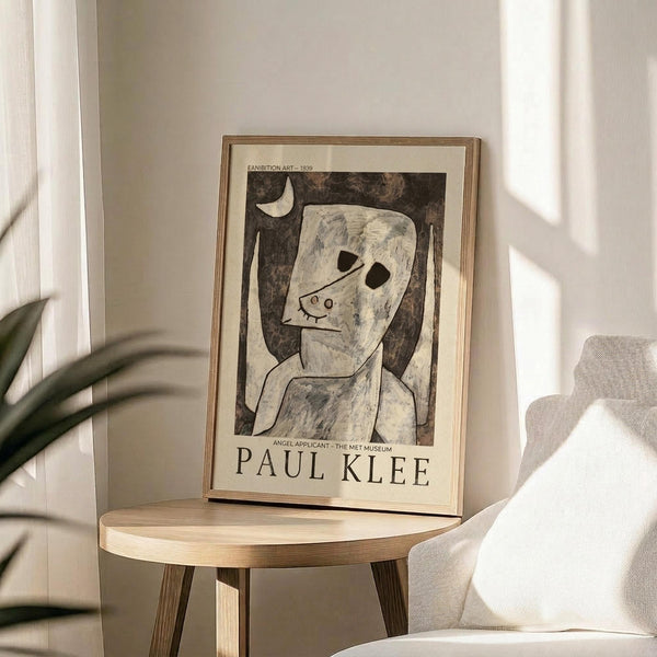 May include: Framed art print featuring Paul Klee's abstract figure with pale face and dark hollow eyes against brown background, displayed on round wooden side table next to beige upholstered chair. Natural sunlight streams across neutral-toned room with plant leaves visible in corner. Poster shows exhibition text referencing The Met Museum and Angel Applicant artwork from 1939.