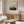 May include: Japanese woodblock-style artwork showing Mount Fuji at sunset with silhouetted buildings and trees in foreground, displayed in thin wooden frame above mid-century modern walnut credenza. Room features cream walls, white curved chair, glass-top oval coffee table, and modern white mushroom lamp. Warm natural lighting creates shadows across the minimalist interior.