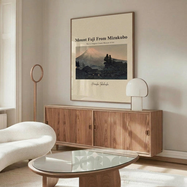 May include: Japanese woodblock-style artwork showing Mount Fuji at sunset with silhouetted buildings and trees in foreground, displayed in thin wooden frame above mid-century modern walnut credenza. Room features cream walls, white curved chair, glass-top oval coffee table, and modern white mushroom lamp. Warm natural lighting creates shadows across the minimalist interior.