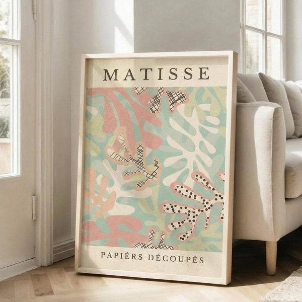 May include: Framed art print in light wood frame leaning against white wall next to beige upholstered sofa. Poster features abstract cut-out style shapes in soft pastels - coral pink, sage green, cream, and yellow - with black crosshatch and dotted patterns. Text reads "MATISSE" at top and "PAPIERS DÉCOUPÉS" at bottom on cream background. Bright natural light streams through tall windows in modern living room setting.