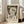 May include: A framed Paul Klee exhibition poster featuring his 1939 "Angel Applicant" artwork displayed on light hardwood floors next to a beige upholstered sofa. The poster shows an abstract figure with angular features and dark hollow eyes against a mottled brown background with a crescent moon, rendered in earth tones. Natural sunlight streams through tall white-framed windows, creating soft shadows on cream-colored walls in this modern living room setting.