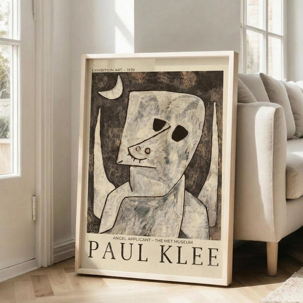May include: A framed Paul Klee exhibition poster featuring his 1939 "Angel Applicant" artwork displayed on light hardwood floors next to a beige upholstered sofa. The poster shows an abstract figure with angular features and dark hollow eyes against a mottled brown background with a crescent moon, rendered in earth tones. Natural sunlight streams through tall white-framed windows, creating soft shadows on cream-colored walls in this modern living room setting.