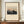 May include: Framed art print featuring Mount Fuji at sunset with pink sky, traditional Japanese village silhouettes in foreground, and dark trees. Cream matting surrounds the artwork in a thin brown wooden frame. Display shows the print leaning against white wainscoting on a mid-century modern walnut credenza, styled with a white ribbed ceramic vase and stacked books.