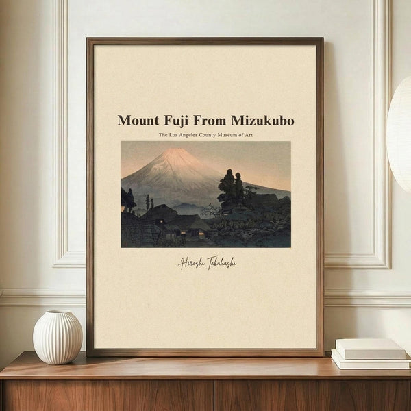 May include: Framed art print featuring Mount Fuji at sunset with pink sky, traditional Japanese village silhouettes in foreground, and dark trees. Cream matting surrounds the artwork in a thin brown wooden frame. Display shows the print leaning against white wainscoting on a mid-century modern walnut credenza, styled with a white ribbed ceramic vase and stacked books.