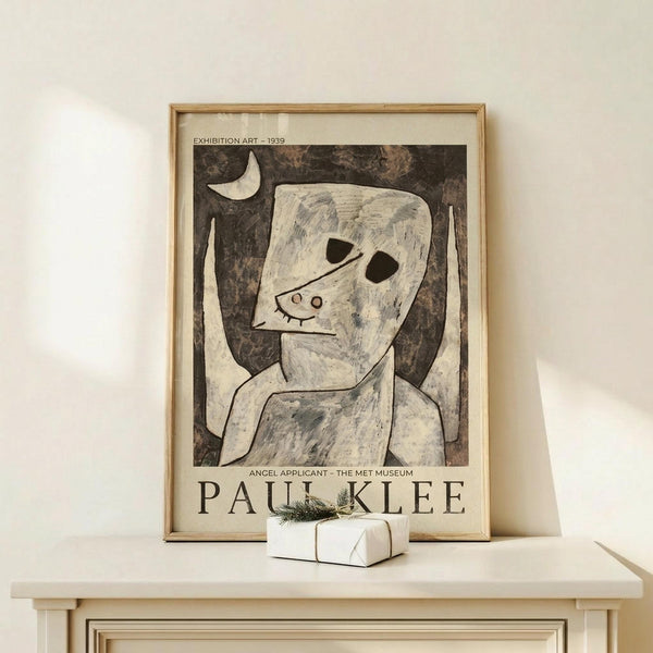 May include: Framed art print featuring Paul Klee's abstract figure with angular white face, dark hollow eyes, and crescent moon against mottled brown background. Print shows "EXHIBITION ART - 1939" and "ANGEL APPLICANT - THE MET MUSEUM" text with "PAUL KLEE" at bottom. Natural wood frame sits on light wooden surface beside small white gift box with green sprig against cream wall.