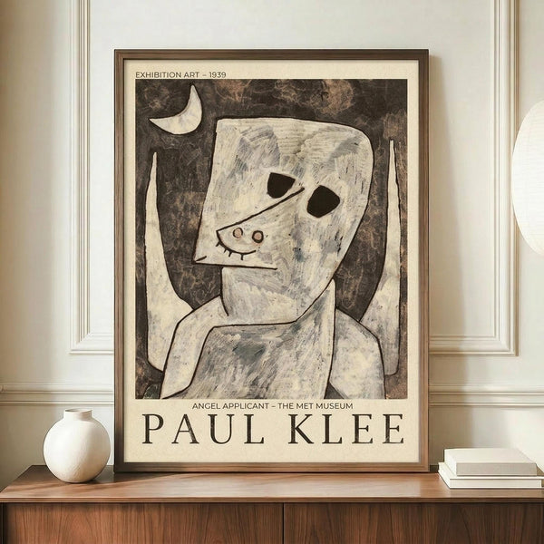 May include: Framed Paul Klee exhibition poster featuring abstract figure with angular white face, dark eye holes, and crescent moon against mottled brown background. Text reads "Exhibition Art - 1939" and "Angel Applicant - The Met Museum" with artist name below. Brown wooden frame sits on mid-century walnut credenza alongside white ceramic vase and stacked books in neutral-toned room with white walls and crown molding.