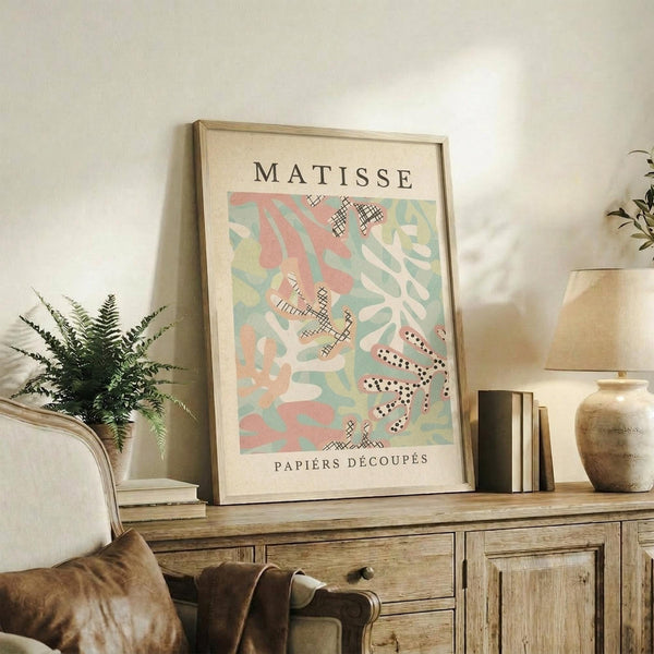 May include: Framed Matisse art print displayed on rustic wooden sideboard in warm-toned living room. Print features abstract cut-out shapes in coral, sage green, and cream with crosshatched and dotted patterns. Natural wood frame complements fern plant, stacked books, ceramic vase, and cream upholstered furniture visible in cozy interior setting.