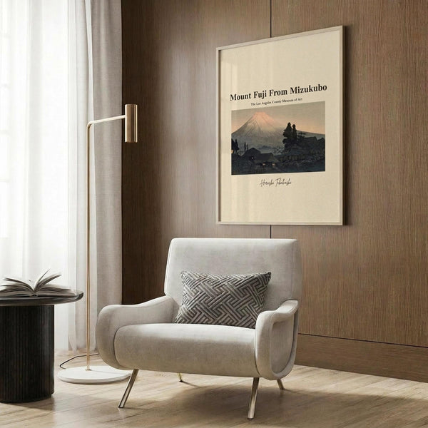 May include: Framed vintage-style art print featuring Mount Fuji at sunset with silhouetted figures in foreground, displayed on wood-paneled wall above modern beige armchair with geometric pillow. Golden floor lamp and dark round side table with open book complete the neutral-toned reading nook with natural lighting from nearby window.