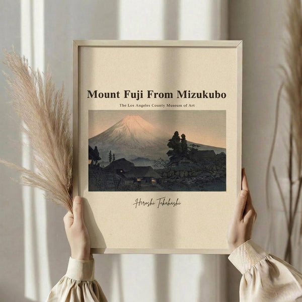 May include: Hands holding a framed poster print showing Mount Fuji with pink sunset sky, dark silhouetted hills and traditional Japanese houses below. Light wood frame on cream mat, held against white curtained window with dried pampas grass visible to left side.