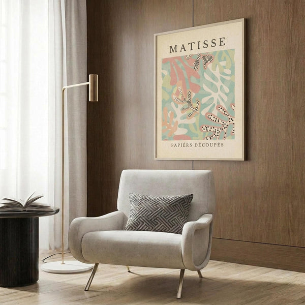 May include: Framed Matisse-style abstract art poster with colorful cut-paper shapes in coral pink, sage green, and cream tones, displayed above a beige upholstered armchair with geometric patterned pillow. Modern brass floor lamp and dark round side table with open book complete the warm, contemporary living room setting with wood-paneled walls.