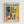 May include: Framed art print in light wood frame on white wall featuring colorful still life scene with leafy plants in red vase, blue and white decorative pottery, bowl of oranges on checkered tablecloth, yellow interior background, styled as vintage exhibition poster with blue "MATISSE" text and "Nice, France" location details