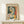 May include: Framed Picasso-style cubist portrait poster featuring a woman with tilted head in geometric forms using green, orange, yellow, and cream tones. Wood frame sits on light wooden surface next to small white wrapped gift with twine. Clean white wall background with soft natural lighting creating gentle shadows.