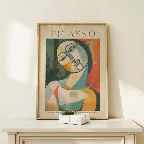 May include: Framed Picasso-style cubist portrait poster featuring a woman with tilted head in geometric forms using green, orange, yellow, and cream tones. Wood frame sits on light wooden surface next to small white wrapped gift with twine. Clean white wall background with soft natural lighting creating gentle shadows.