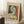 May include: Framed Picasso-style cubist portrait poster leaning against white wall corner on textured white rug. Abstract woman's face in geometric shapes with green, orange, yellow, and blue colors. Light wood frame, cream matting. Coffee cup on wooden block nearby, beige blanket visible. Natural window light creates soft shadows.