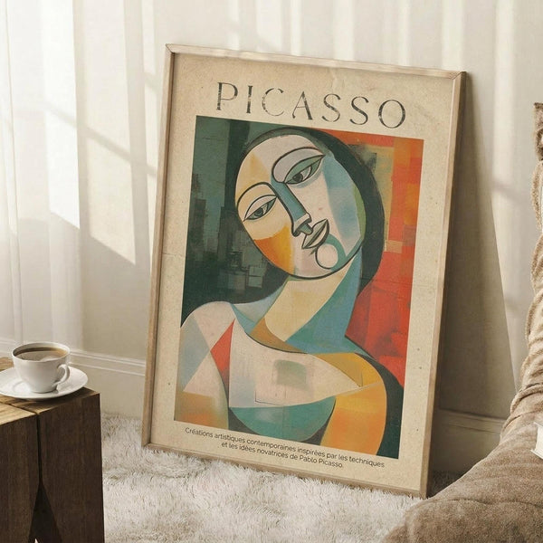 May include: Framed Picasso-style cubist portrait poster leaning against white wall corner on textured white rug. Abstract woman's face in geometric shapes with green, orange, yellow, and blue colors. Light wood frame, cream matting. Coffee cup on wooden block nearby, beige blanket visible. Natural window light creates soft shadows.