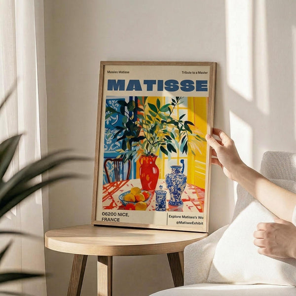 May include: Framed vintage-style art poster featuring Matisse's colorful still life with plants in vases, fruit bowl, and decorative objects in blues, yellows, and reds. Wooden frame sits on round wooden side table next to beige upholstered chair. Person's hands visible adjusting the frame. Soft natural lighting creates shadows on white wall background with plant leaves visible in corner.