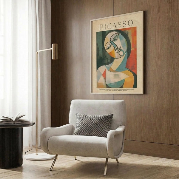 May include: A framed Picasso-style cubist portrait poster featuring a stylized woman's face in geometric shapes with green, orange, yellow and cream colors, displayed on a wood-paneled wall above a beige upholstered armchair with geometric patterned pillow, next to a brass floor lamp and dark round side table with open book.