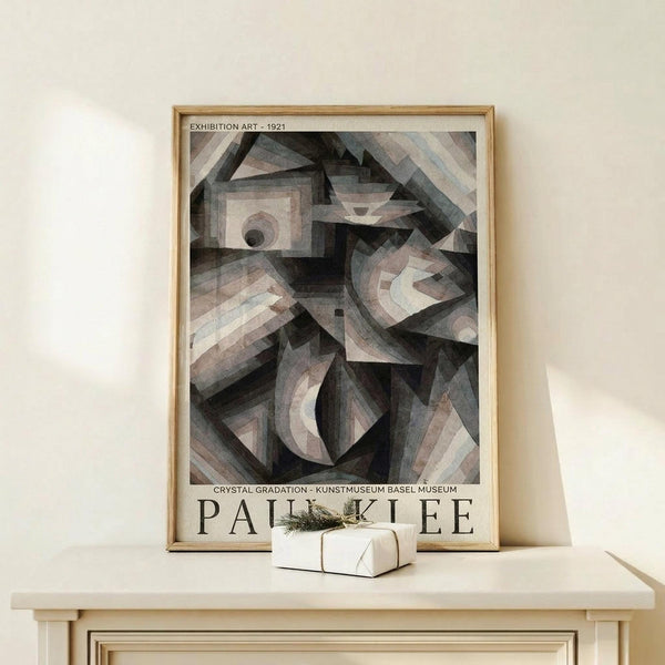 May include: Framed poster print featuring Paul Klee's abstract geometric artwork in muted tones of gray, beige, and soft pastels. The cubist-style composition shows overlapping angular shapes, curves, and forms creating depth and movement. Text reads "Exhibition Art - 1921" at top and "Crystal Gradation - Kunstmuseum Basel Museum" with "Paul Klee" at bottom. Natural wood frame rests on light wooden surface against cream wall, with small wrapped gift beside it.