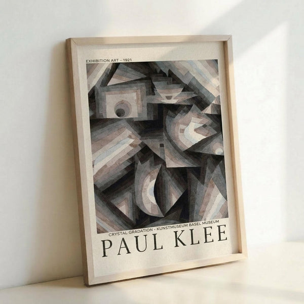 May include: Framed exhibition poster featuring Paul Klee's abstract geometric artwork from 1921 in muted grays, browns and beiges. The cubist-style composition shows angular, crystalline forms with circular and triangular shapes creating depth and movement. Text reads "Exhibition Art - 1921" at top and "Crystal Gradation - Kunstmuseum Basel Museum" and "Paul Klee" at bottom. Displayed in light wood frame leaning against white wall.