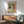May include: Framed Picasso-style abstract portrait poster showing stylized figure with geometric face in green, orange, and cream tones, displayed above mid-century modern wooden sideboard with table lamp, next to sage green upholstered armchair with wooden frame in sunlit room with white walls