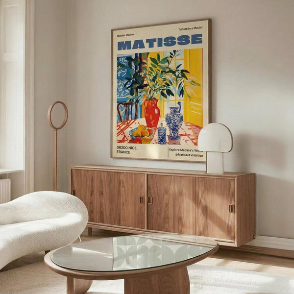 May include: Colorful Matisse exhibition poster in wooden frame hanging above walnut credenza in modern living room. Poster features vibrant still life with plants, vases, and decorative objects in blues, yellows, and reds. White sculptural chair and glass-top coffee table visible in foreground with floor lamp beside credenza.