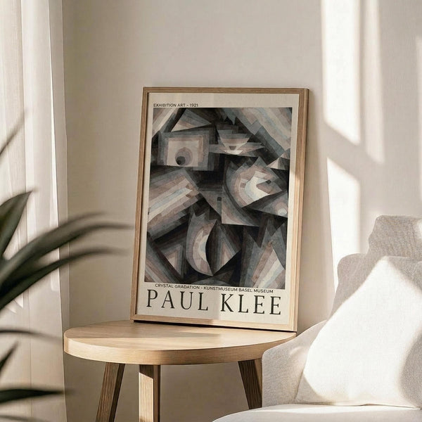 May include: Framed exhibition poster featuring Paul Klee's abstract geometric artwork with overlapping circles, triangles and angular shapes in muted grays, blues and earth tones. The poster sits on a light wood round side table next to a beige upholstered chair, with a potted plant visible in soft natural lighting casting shadows on cream walls.