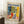 May include: Framed vintage-style exhibition poster featuring Matisse's colorful still life with leafy plants in red vase, blue and white ceramic vessels, and fruit bowl on checkered tablecloth against yellow window light. Poster leans against white wall next to beige armchair and wooden side table with white coffee cup, displayed on textured cream rug in bright room setting.