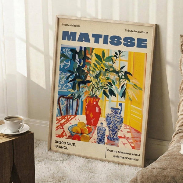 May include: Framed vintage-style exhibition poster featuring Matisse's colorful still life with leafy plants in red vase, blue and white ceramic vessels, and fruit bowl on checkered tablecloth against yellow window light. Poster leans against white wall next to beige armchair and wooden side table with white coffee cup, displayed on textured cream rug in bright room setting.