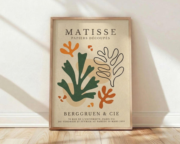 May include: Framed art print in light wood frame displaying abstract cut-out shapes in orange, green, and beige on cream background with French text. Print features organic coral-like forms and flowing botanical elements. Frame sits on light wood floor against white wall with soft natural lighting creating diagonal shadows.