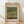 May include: Framed exhibition poster featuring Claude Monet's Japanese Footbridge painting from 1899, showing a curved blue-green bridge over a lily pond with water lilies and lush garden vegetation. The poster has cream-colored matting with elegant typography and is displayed in a light wood frame, leaning against a white wall on light wooden flooring with natural window light creating soft shadows.