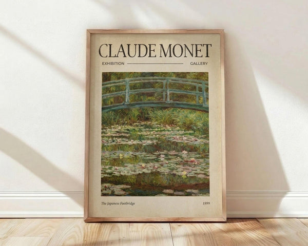 May include: Framed exhibition poster featuring Claude Monet's Japanese Footbridge painting from 1899, showing a curved blue-green bridge over a lily pond with water lilies and lush garden vegetation. The poster has cream-colored matting with elegant typography and is displayed in a light wood frame, leaning against a white wall on light wooden flooring with natural window light creating soft shadows.