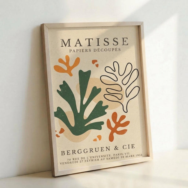 May include: Framed vintage-style art print featuring Matisse paper cutout designs in green, orange, and beige organic shapes resembling coral or seaweed forms. Cream background with "Berggruen & Cie" gallery text and Paris exhibition details. Light wooden frame leaning against white wall on light surface.