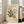 May include: Framed art print in light wood frame leaning against white wall next to beige upholstered chair. Print features cream background with abstract cutout-style shapes in green, orange, and beige resembling seaweed or coral forms. Black text at top and bottom. Sunlight streams through nearby windows onto hardwood floor.