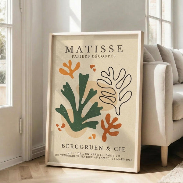 May include: Framed art print in light wood frame leaning against white wall next to beige upholstered chair. Print features cream background with abstract cutout-style shapes in green, orange, and beige resembling seaweed or coral forms. Black text at top and bottom. Sunlight streams through nearby windows onto hardwood floor.