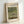 May include: Framed exhibition poster featuring Claude Monet's Japanese Footbridge painting from 1899, showing a curved blue-green bridge over a pond with water lilies and lush vegetation. The poster has clean typography with "CLAUDE MONET EXHIBITION GALLERY" at the top and artwork details below. Light wooden frame sits against white wall, casting subtle shadows.