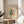 May include: Framed art print in thin wooden frame displaying abstract cut-out style botanical shapes in green, orange, and cream colors with French text, positioned on round wooden side table next to beige upholstered chair and plant leaves, with natural sunlight casting shadows on neutral wall.